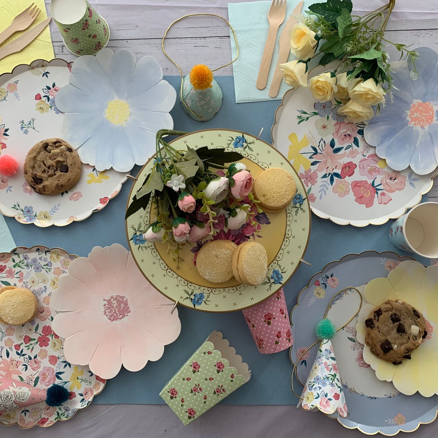 Children's tea party setup with floral plates, cookies, and party hats on a blue tablecloth.